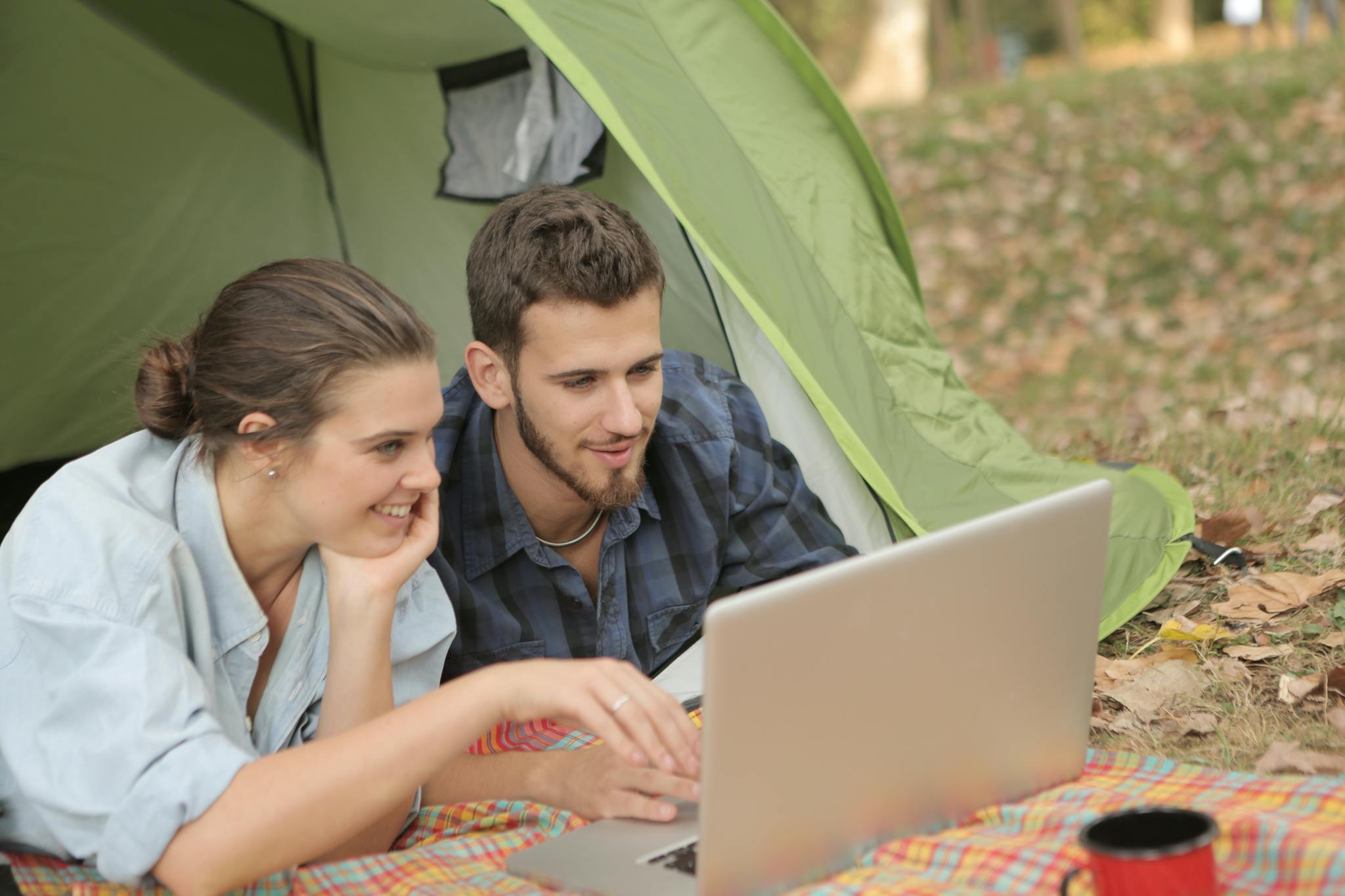 Cheerful female and bearded male using laptop with touchpad while resting in tent at campsite